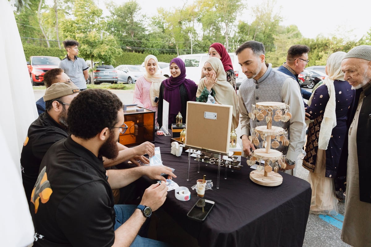 Community vendor at outdoor market event demonstrating products to diverse group of visitors at a table display