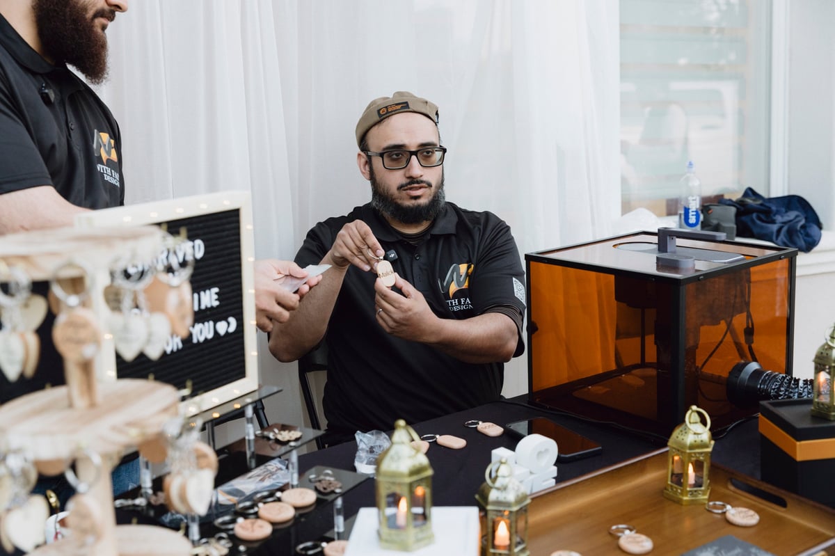 Man with glasses and beard in black shirt working at desk with 3D printer and jewelry-making tools in studio setting