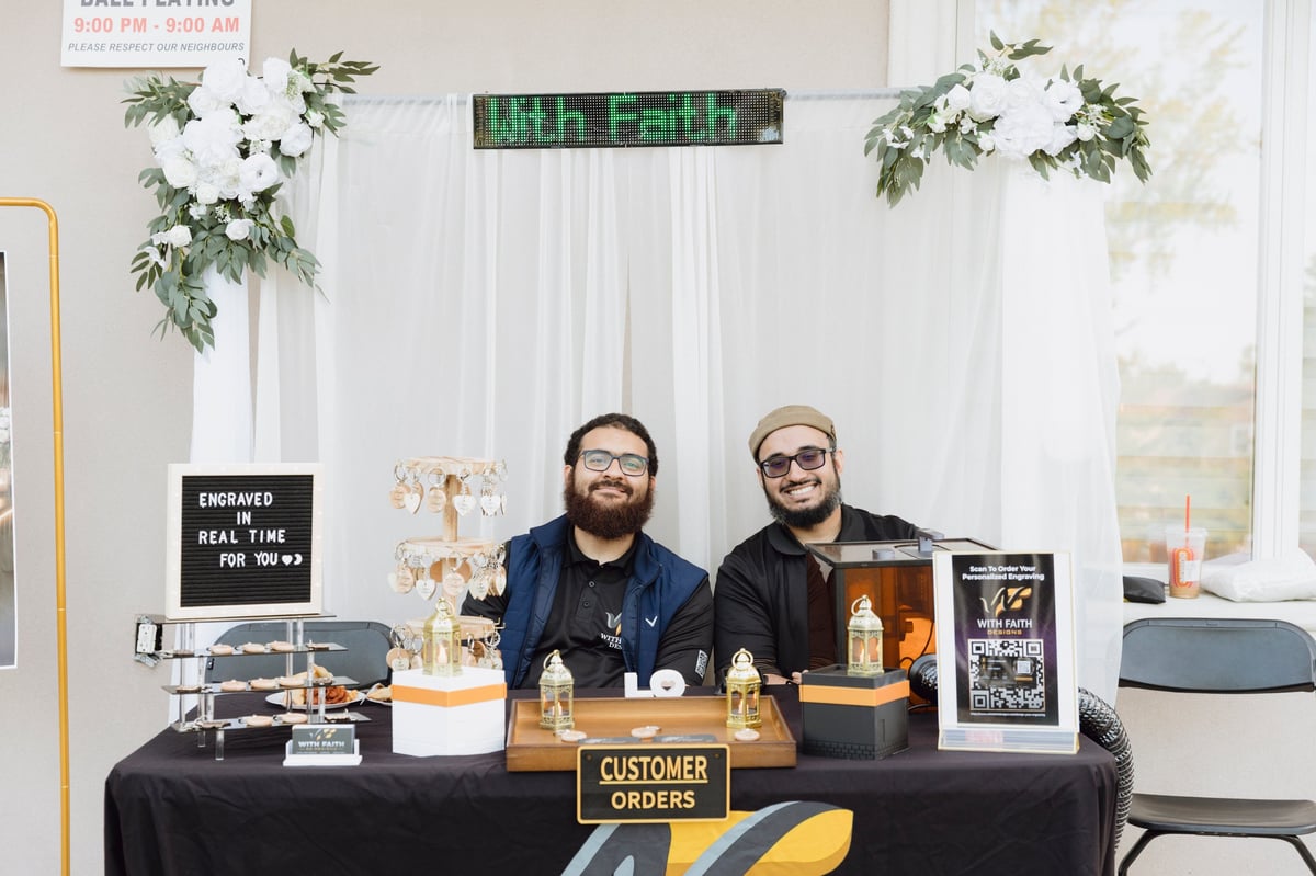 Two men wearing glasses sit behind a vendor table decorated with white curtains and greenery at an event.