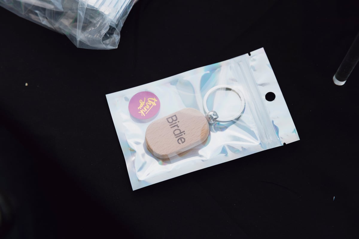 Overhead view of a beige keychain with blue branding on a white patterned pouch, accompanied by a pink circular sticker on a black surface